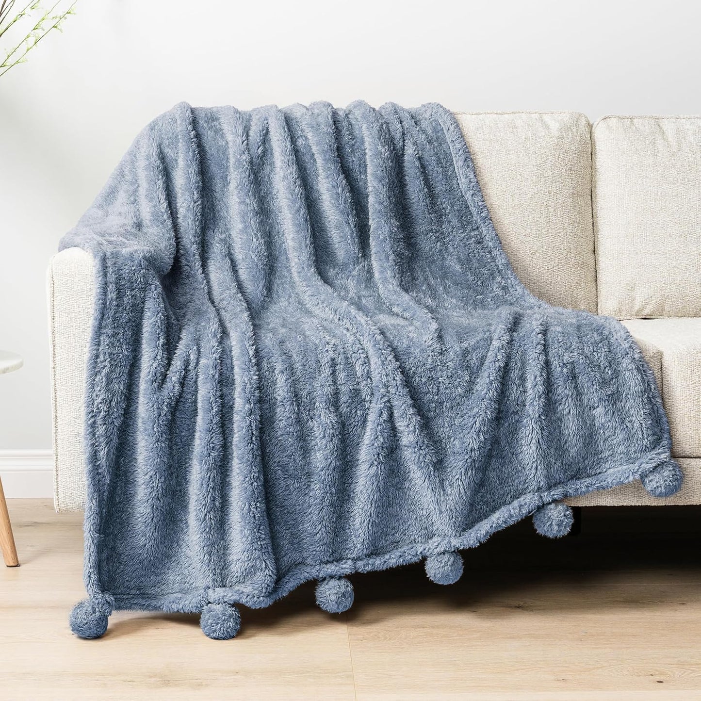 Blue fluffy blanket with pom-pom edges draped over a beige sofa.