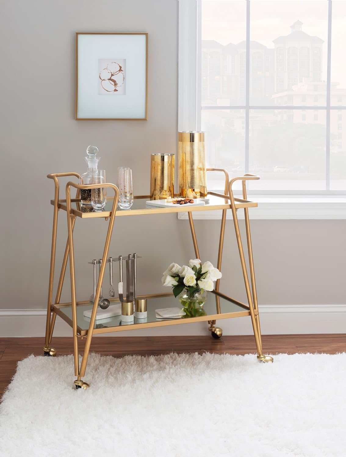 Gold bar cart with decorative items in a room with a window and framed picture on the wall.